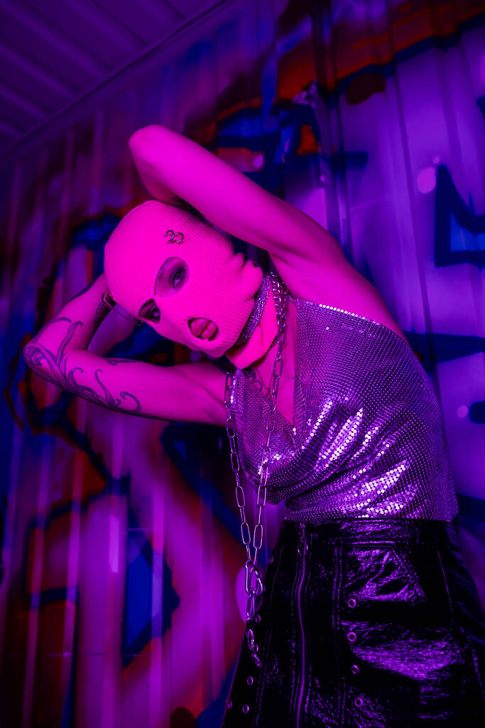 vista de ángulo bajo de la mujer provocativa en pasamontañas y la parte superior metálica mirando a la cámara en la luz púrpura cerca de graffiti colorido - Foto, imagen