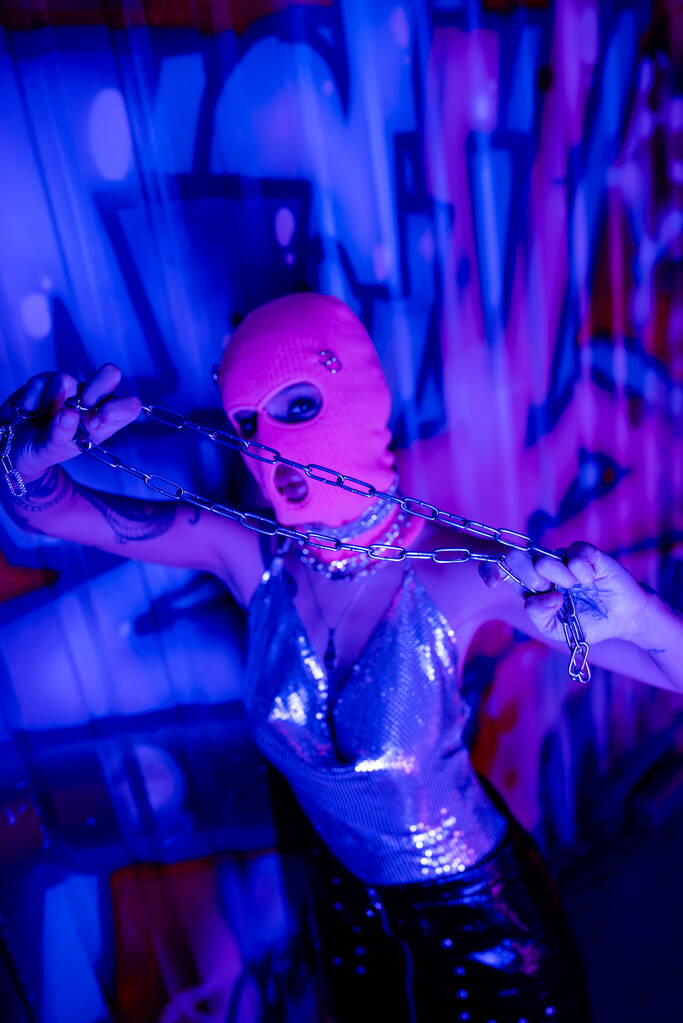 высокий угол обзора провокационной женщины в балаклаве, смотрящей в камеру, позируя с серебряной цепью рядом с граффити в синем неоновом свете - Фото, изображение