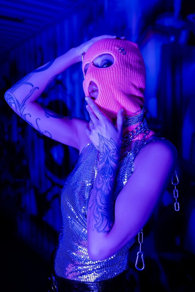 mujer tatuada en la parte superior brillante y pasamontañas cogidas de la mano cerca de la cara mientras mira a la cámara en luz azul y púrpura - Foto, imagen