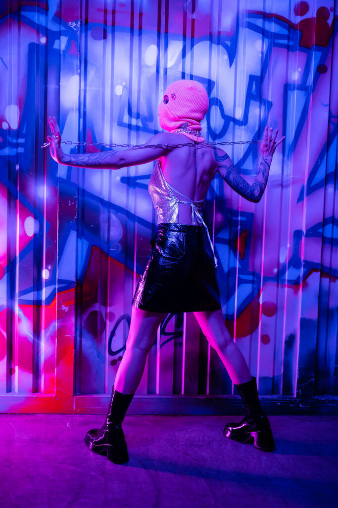 vista trasera de mujer provocativa en botas negras y pasamontañas posando con cadena de plata cerca de coloridos graffiti en luz de neón azul - Foto, imagen