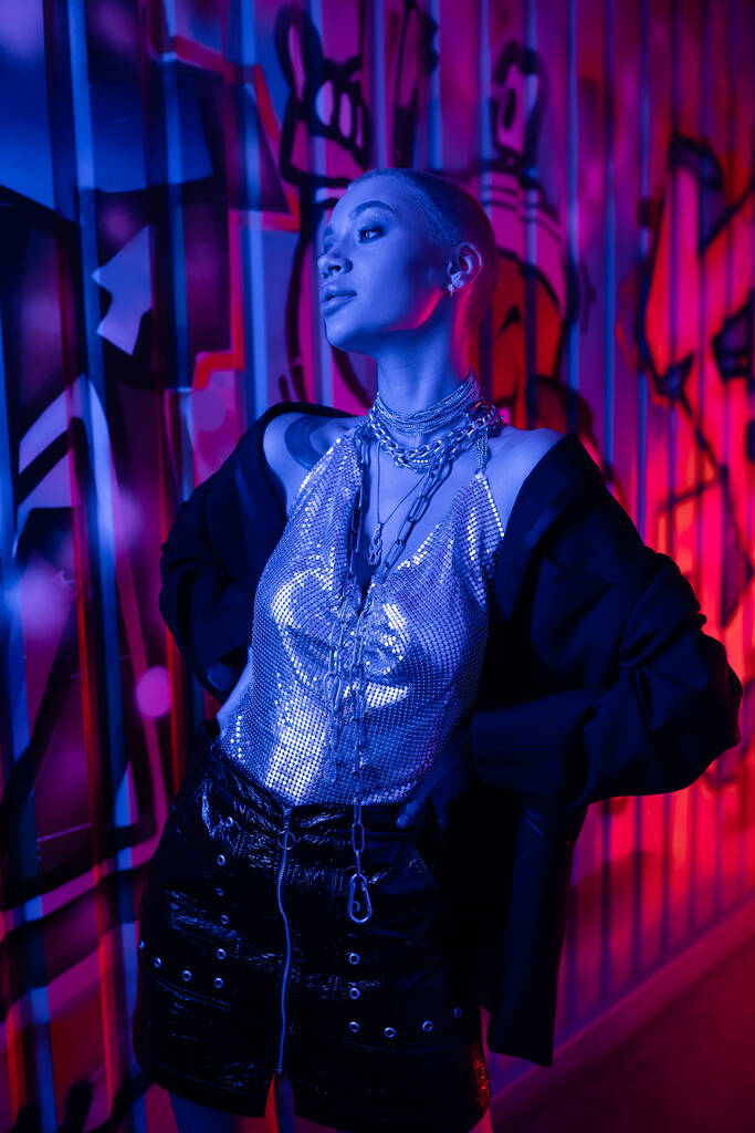 блискуча жінка в металевому верхньому і срібному ланцюжках шиї позує руками на талії біля барвистого графіті в блакитному неоновому світлі
 - Фото, зображення