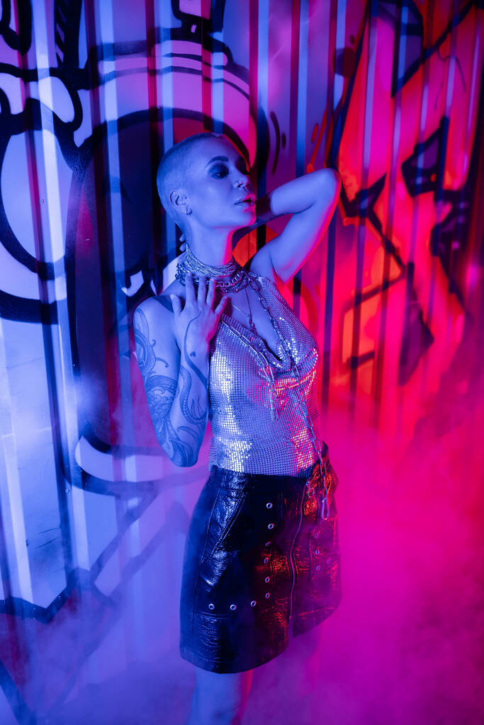 appassionata donna tatuata in top metallico e gonna in pelle nera in piedi vicino a graffiti colorati in luce blu e rosa con fumo - Foto, immagini