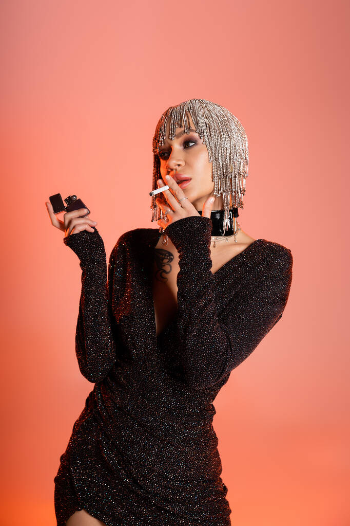 金属製のヘッドウェアを着た魅惑的な女性と黒のルレックスのドレス喫煙タバコサンゴピンクの背景 - 写真・画像
