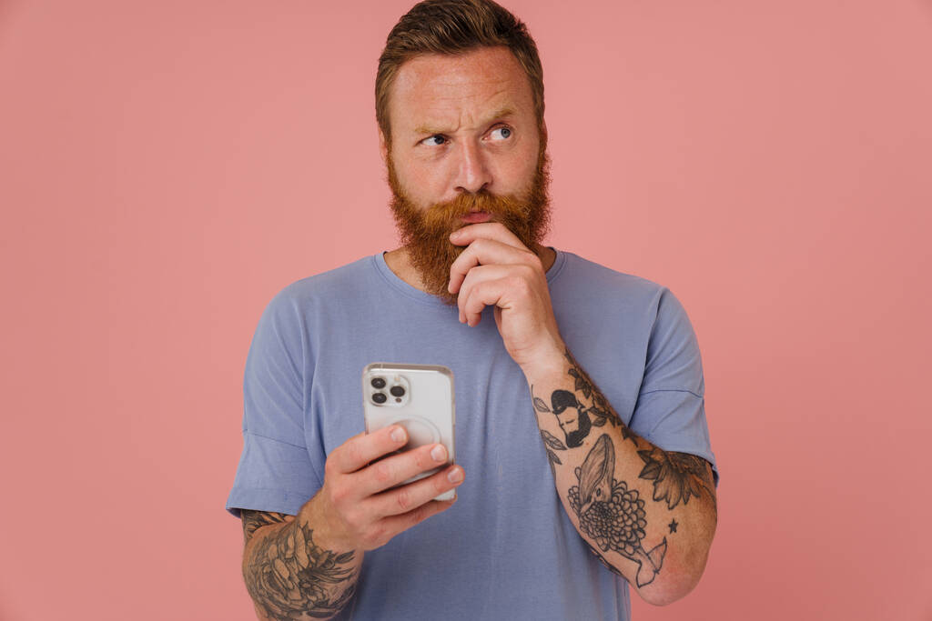 Portré felnőtt szakállas tetovált jóképű figyelmes férfi néz félre, és tartja a szakállát, miközben áll felett elszigetelt rózsaszín háttér - Fotó, kép