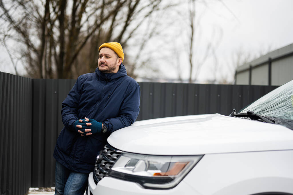 Mies kuljettaja käyttää takki ja keltainen hattu vastaan nojasi huppu amerikkalainen maasturi auton kylmällä säällä. - Valokuva, kuva