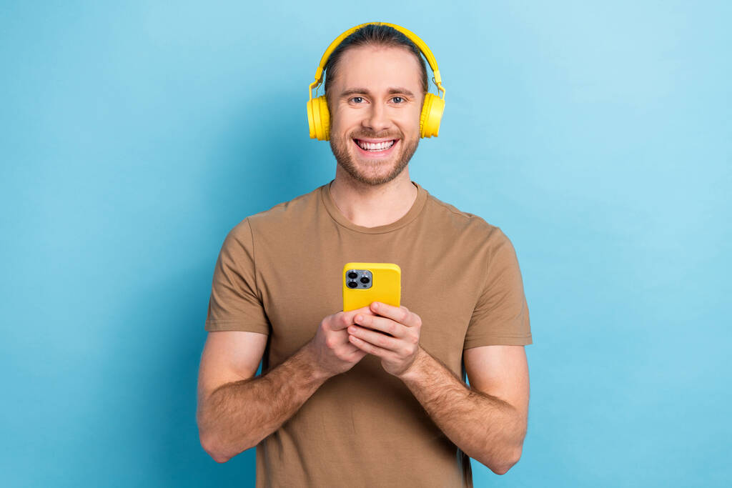 Fotografie pozitivní veselý chlap oblečený béžové tričko sluchátka poslech hudba moderní gadget izolované modré barvy pozadí. - Fotografie, Obrázek