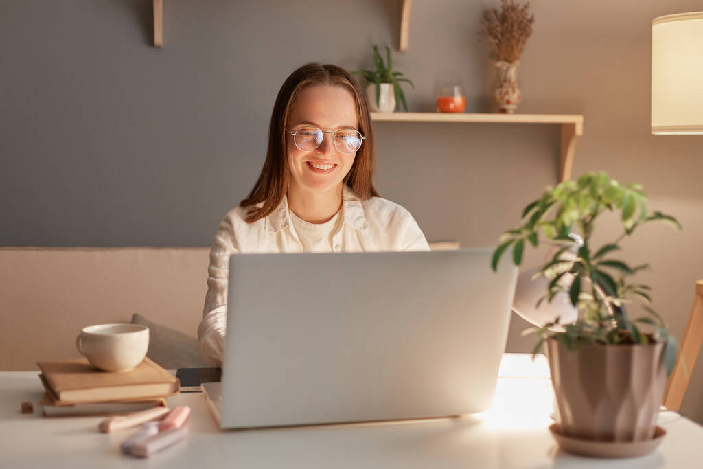 Plan horizontal d'une belle femme pigiste ou étudiante portant une chemise blanche et des lunettes, assise au bureau à la maison le soir, travaillant sur un ordinateur portable, regardant l'écran - Photo, image