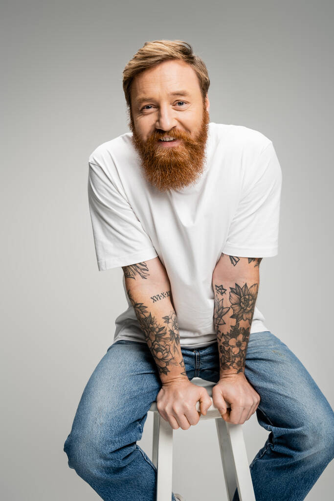 Веселий татуйований чоловік у повсякденному одязі сидить на стільці ізольовано на сірому
  - Фото, зображення