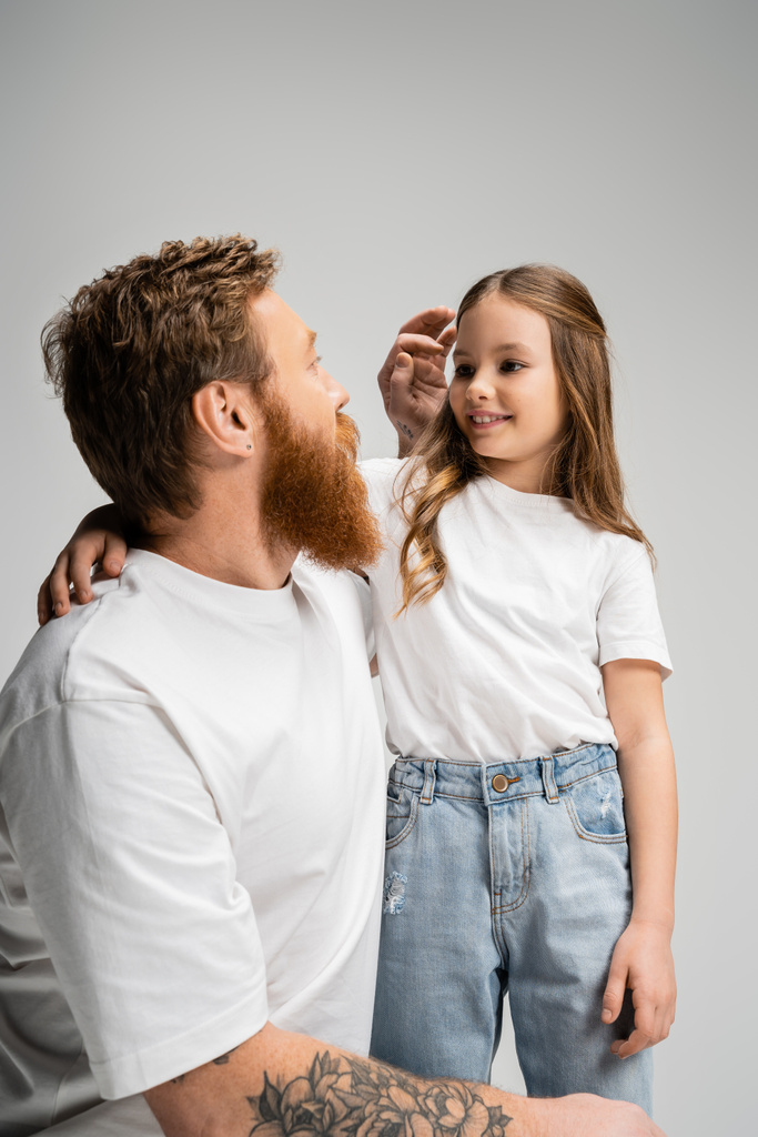 Γενειοφόρος πατέρας προσαρμόζει τα μαλλιά της χαμογελαστής κόρης απομονώνονται σε γκρι  - Φωτογραφία, εικόνα
