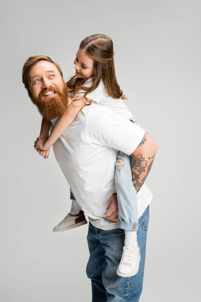 Веселый ребенок, сидящий на татуированном отце с бородой, изолированной на сером  - Фото, изображение