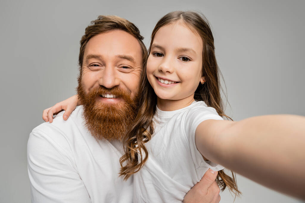 Χαμογελώντας preteen παιδί αγκαλιάζει γενειοφόρος μπαμπάς σε λευκό t-shirt απομονωμένο σε γκρι  - Φωτογραφία, εικόνα