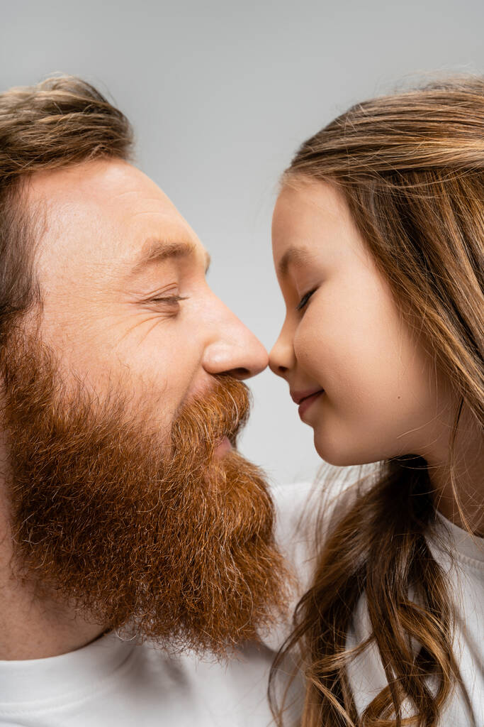 Vista lateral do pai barbudo e da filha pré-adolescente de pé nariz a nariz isolado no cinza  - Foto, Imagem