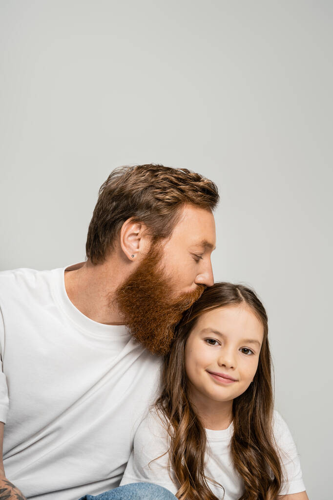 Bärtiger Mann im weißen T-Shirt küsst lächelnde Tochter isoliert auf grau   - Foto, Bild