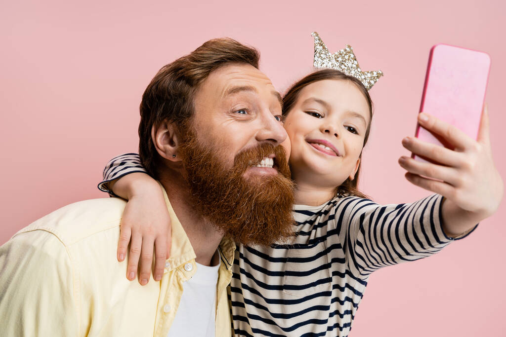 Chica sonriente con diadema de corona abrazando a papá barbudo mientras toma selfie aislado en rosa   - Foto, imagen