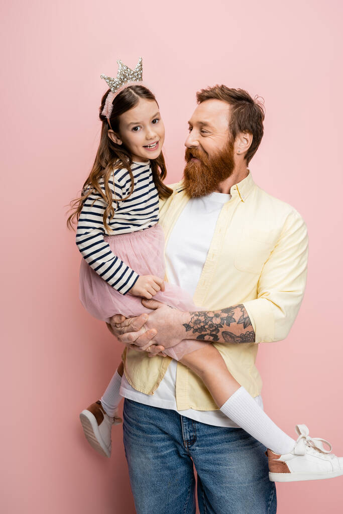 Tätowierter Mann hält Tochter mit Krone-Stirnband auf rosa Hintergrund  - Foto, Bild