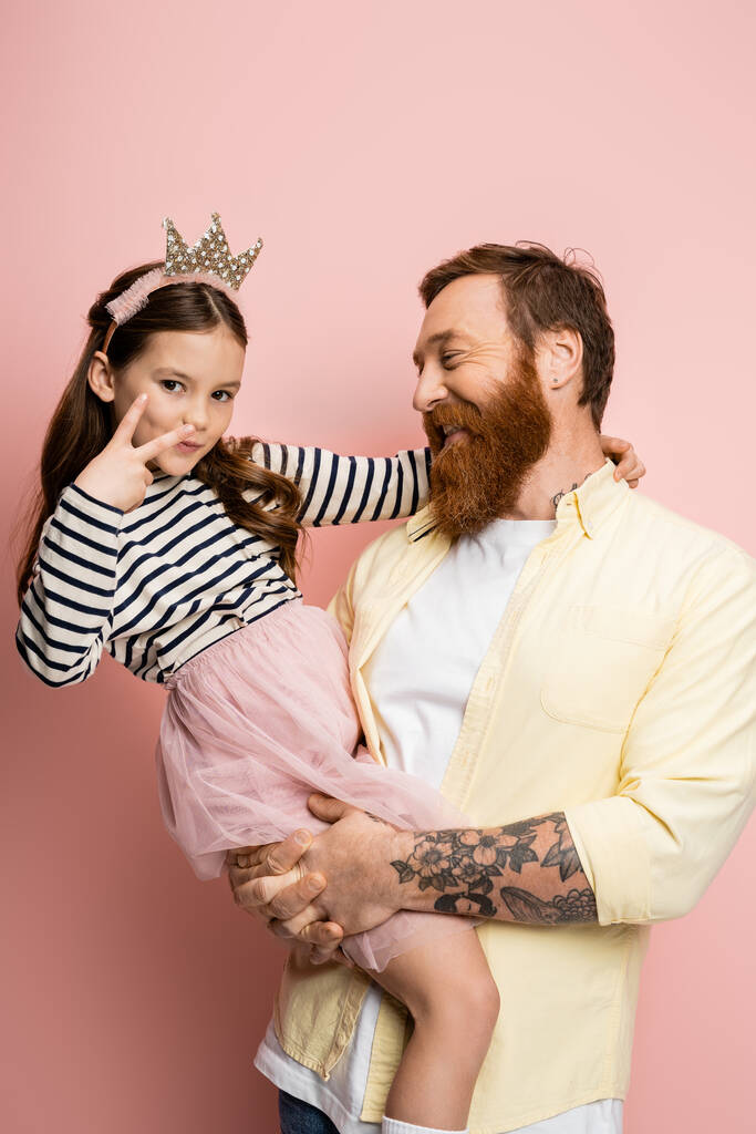Nadšený muž drží preteen dcera s korunou čelenka ukazující mír znamení na růžovém pozadí  - Fotografie, Obrázek