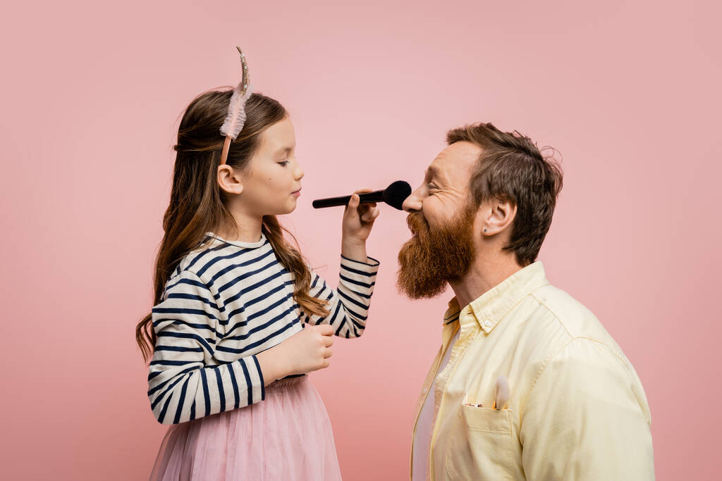 Seitenansicht des Kindes in Krone Stirnband hält Make-up-Pinsel in der Nähe Vater isoliert auf rosa   - Foto, Bild