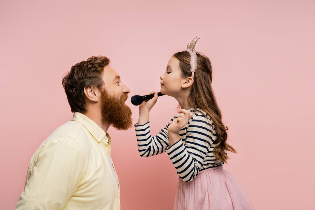 Seitenansicht des Mädchens in Krone Stirnband hält Make-up-Pinsel in der Nähe fröhlichen Vater isoliert auf rosa   - Foto, Bild