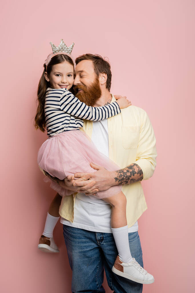 Tattooed man holding cheerful daughter with crown headband on pink background  - Photo, Image