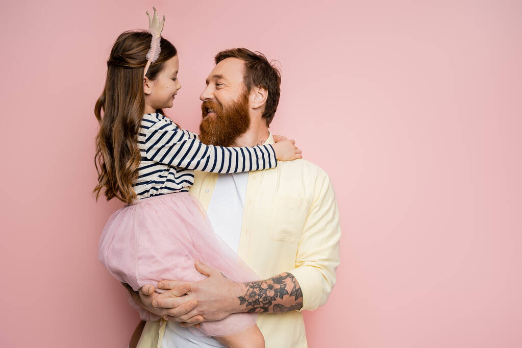 Hombre tatuado positivo sosteniendo hija con diadema de corona sobre fondo rosa  - Foto, imagen