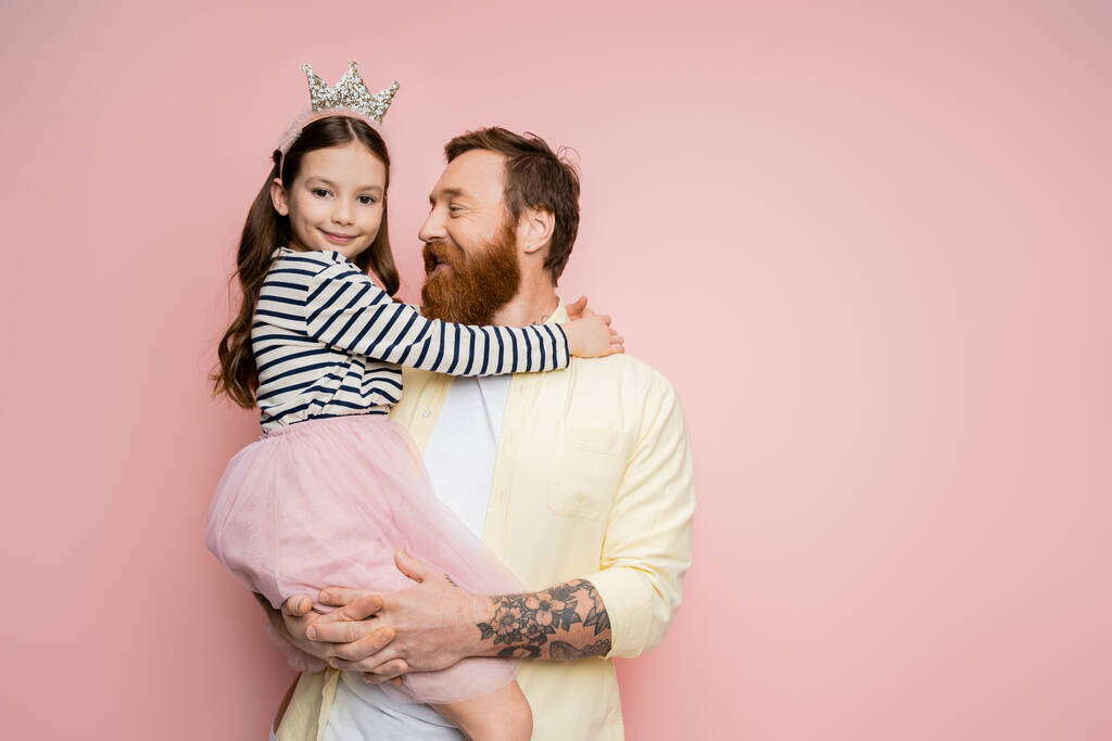 Χαρούμενος γενειοφόρος πατέρας κρατώντας κόρη με κορδέλα στο κεφάλι σε ροζ φόντο  - Φωτογραφία, εικόνα