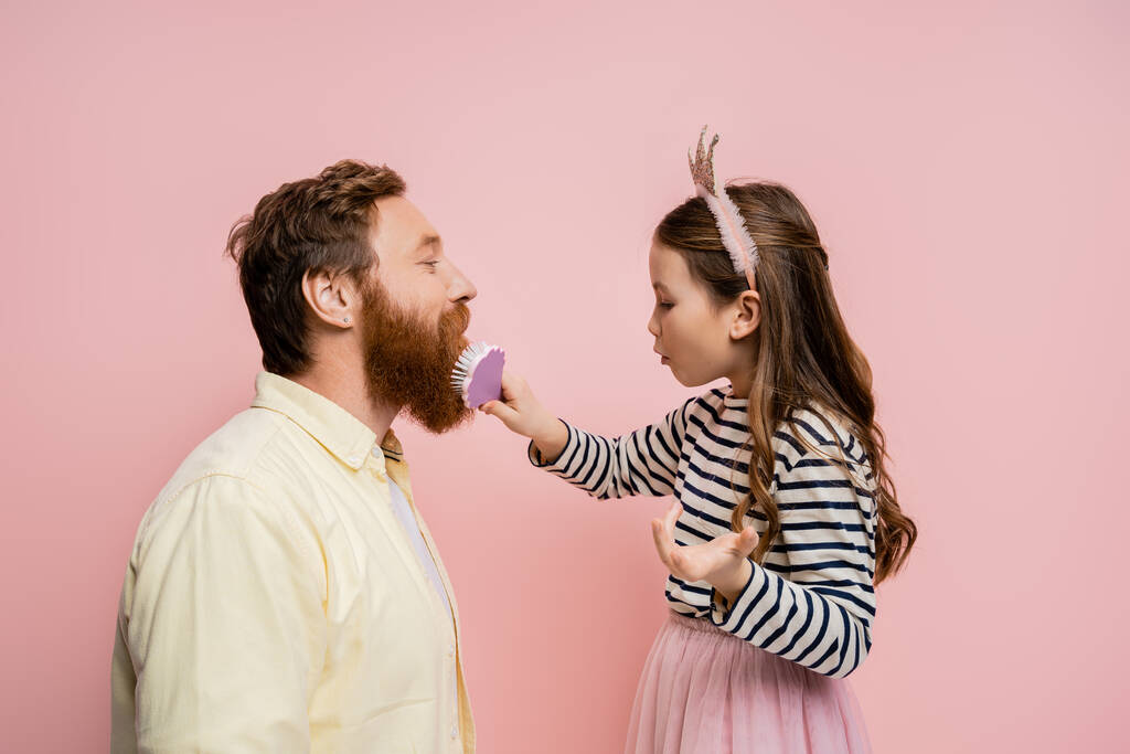 Side view of preteen girl with crown headband brushing beard of father isolated on pink   - Photo, Image