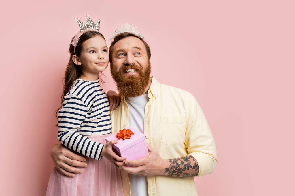 Positive Vater und Tochter mit Krone Stirnbänder halten Geschenk und schauen weg auf rosa Hintergrund  - Foto, Bild