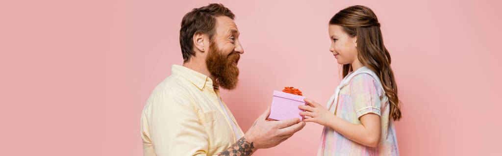 Боковой вид татуированного отца, делающего подарок дочери на розовом баннере  - Фото, изображение
