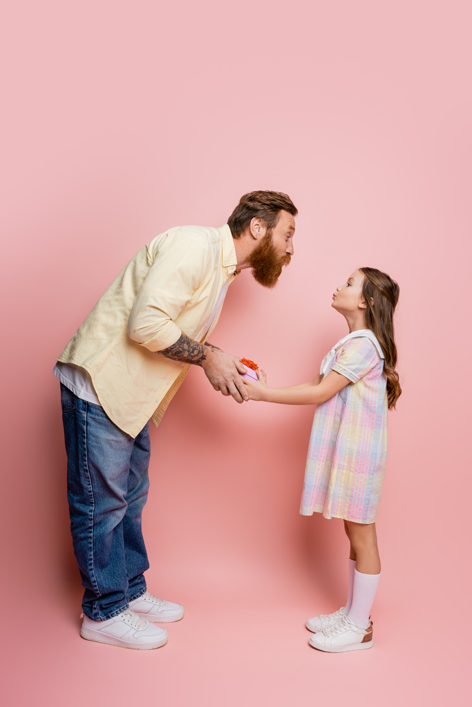 Vista lateral do homem amuando os lábios enquanto dá caixa de presente para a filha no fundo rosa  - Foto, Imagem