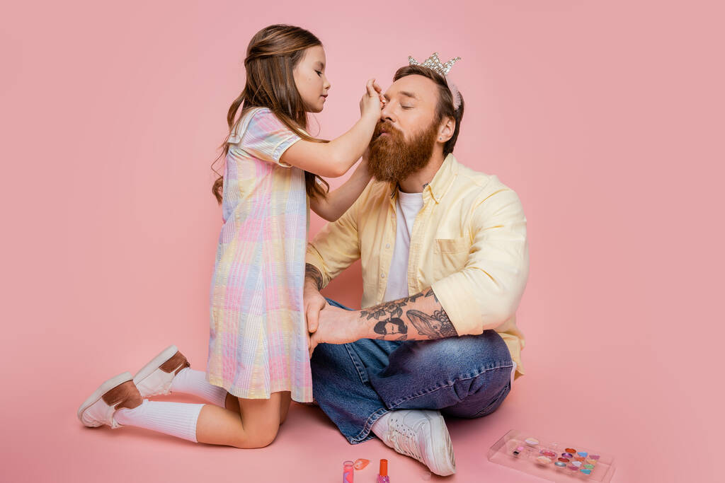 Chica preadolescente aplicando cosmética decorativa en papá tatuado con diadema de corona sobre fondo rosa  - Foto, imagen