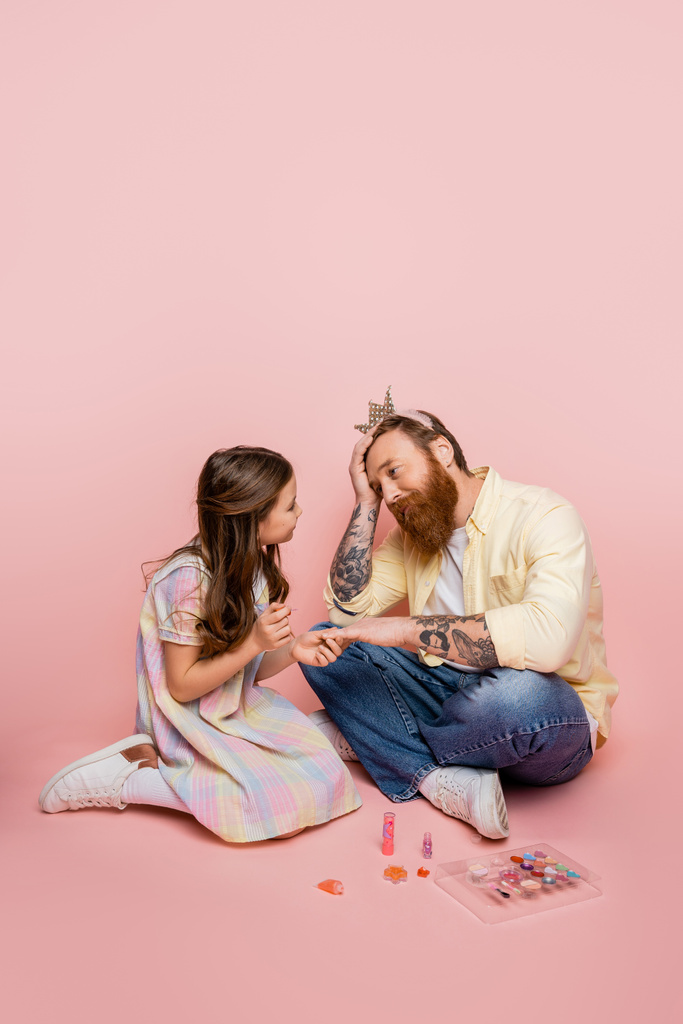Preteen girl holding nail polish near hand of tired father with crown headband on pink background  - Photo, Image