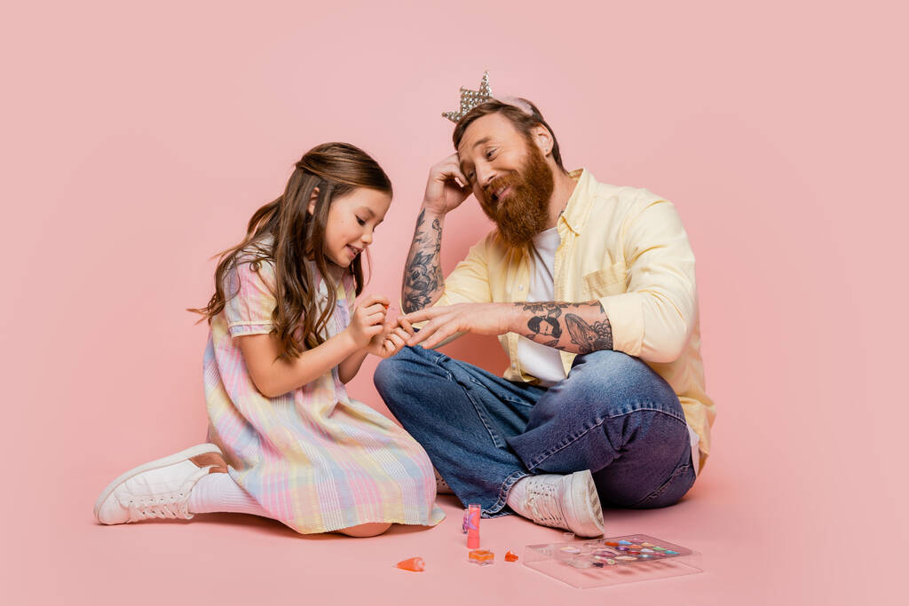 Fröhliches Mädchen, das Nagellack auf der Hand des tätowierten Papas mit Krone Stirnband auf rosa Hintergrund aufträgt  - Foto, Bild