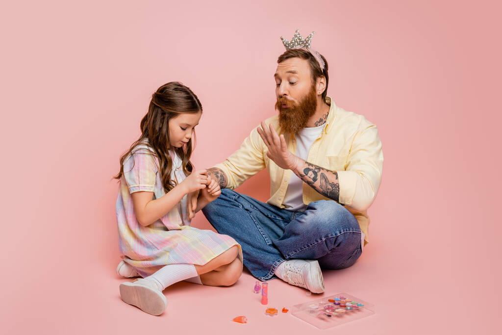 Hombre con corona en la cabeza soplando en la mano cerca de la hija con esmalte de uñas sobre fondo rosa  - Foto, imagen