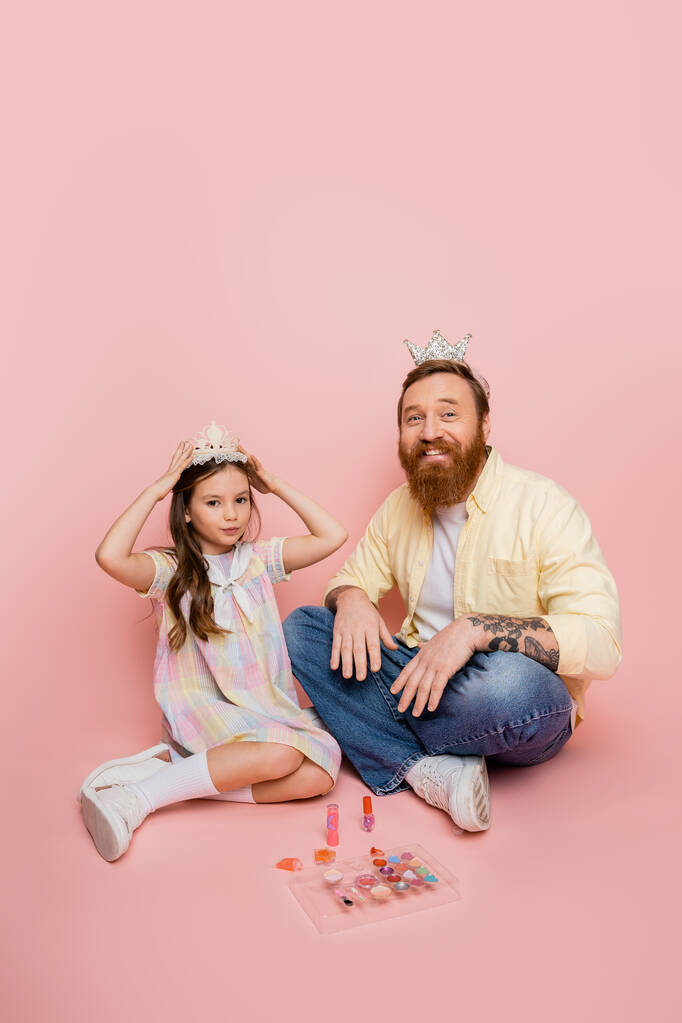Positiver bärtiger Mann blickt in die Kamera nahe Tochter mit Krone und dekorativer Kosmetik auf rosa Hintergrund  - Foto, Bild