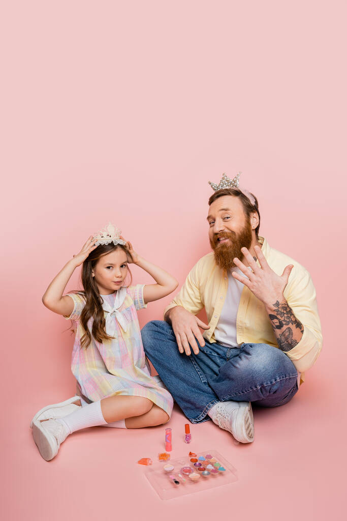 niña preadolescente con corona cerca de padre barbudo y cosméticos decorativos sobre fondo rosa  - Foto, imagen