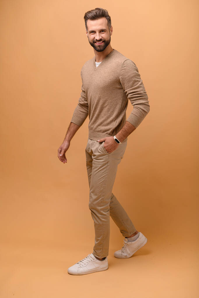 Full length portrait of handsome bearded man in beige pullover is holding hand in pockets and walking, guy in stylish eyeglasses looking at the camera with pleasant smile, isolated studio shot - Photo, Image