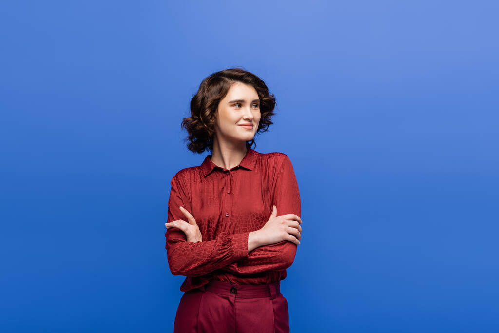 cheerful woman with short curly hair standing with folded arms isolated on blue  - Photo, Image