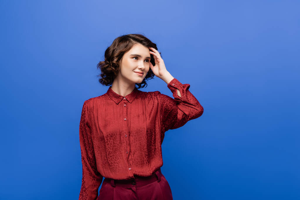 pensive language teacher in red blouse smiling while looking away isolated on blue  - Photo, Image