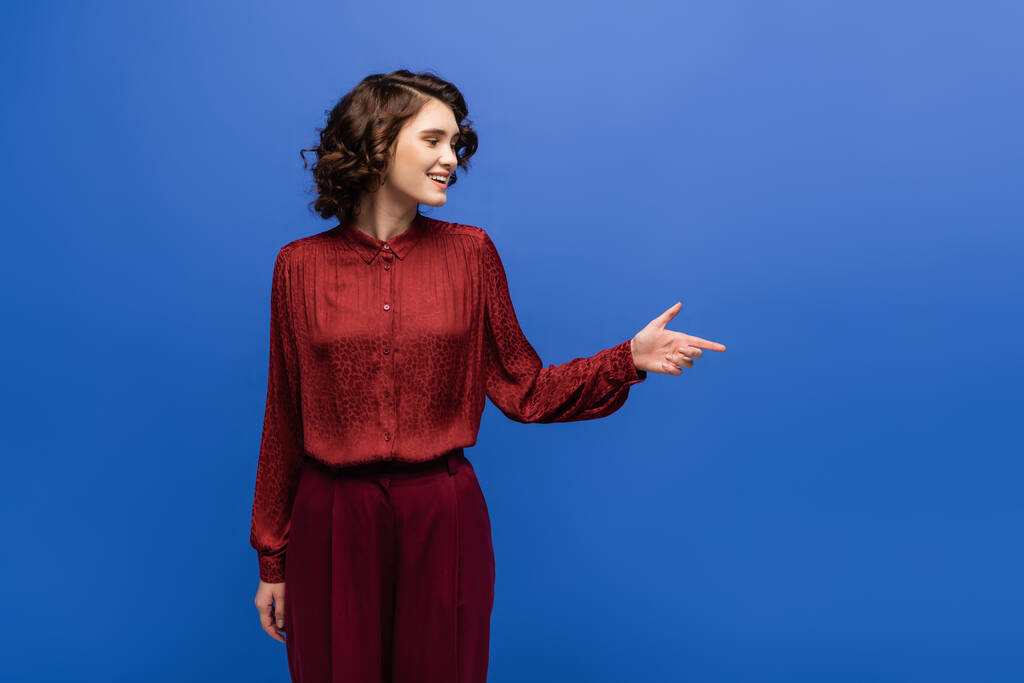 cheerful language teacher in maroon color outfit smiling while pointing with finger isolated on blue  - Photo, Image