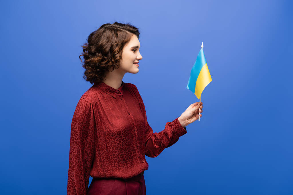 vista laterale di felice insegnante di lingua in possesso di bandiera dell'Ucraina isolato su blu  - Foto, immagini