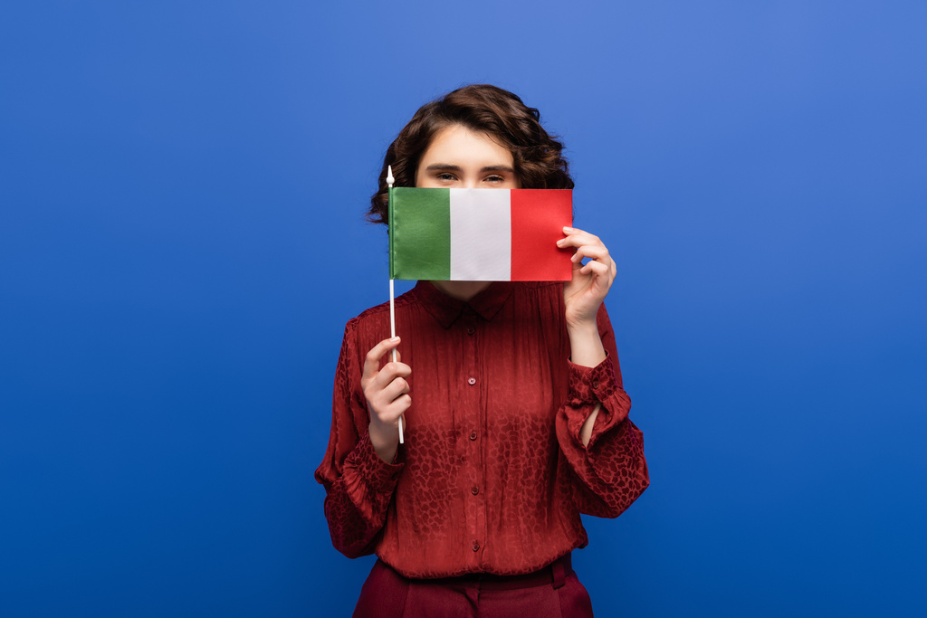 kręcony nauczyciel języka zakrywający twarz trzymając flagę Włoch i patrząc w kamerę odizolowaną na niebiesko  - Zdjęcie, obraz