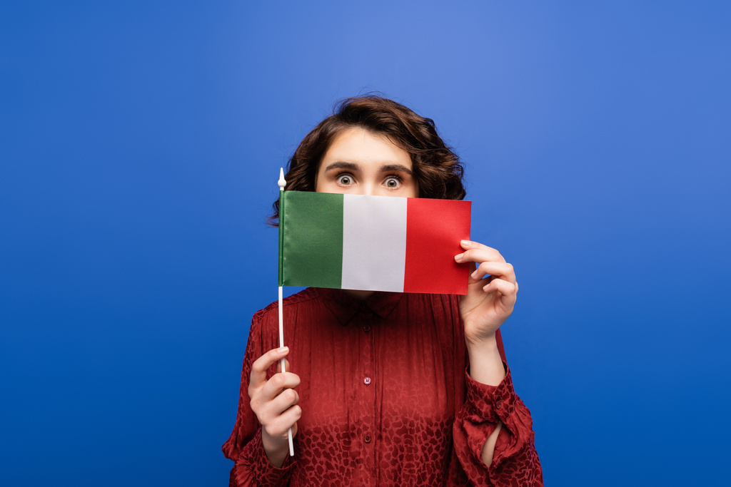 giovane donna riccia che copre il viso tenendo la bandiera d'Italia e guardando la fotocamera isolata sul blu  - Foto, immagini