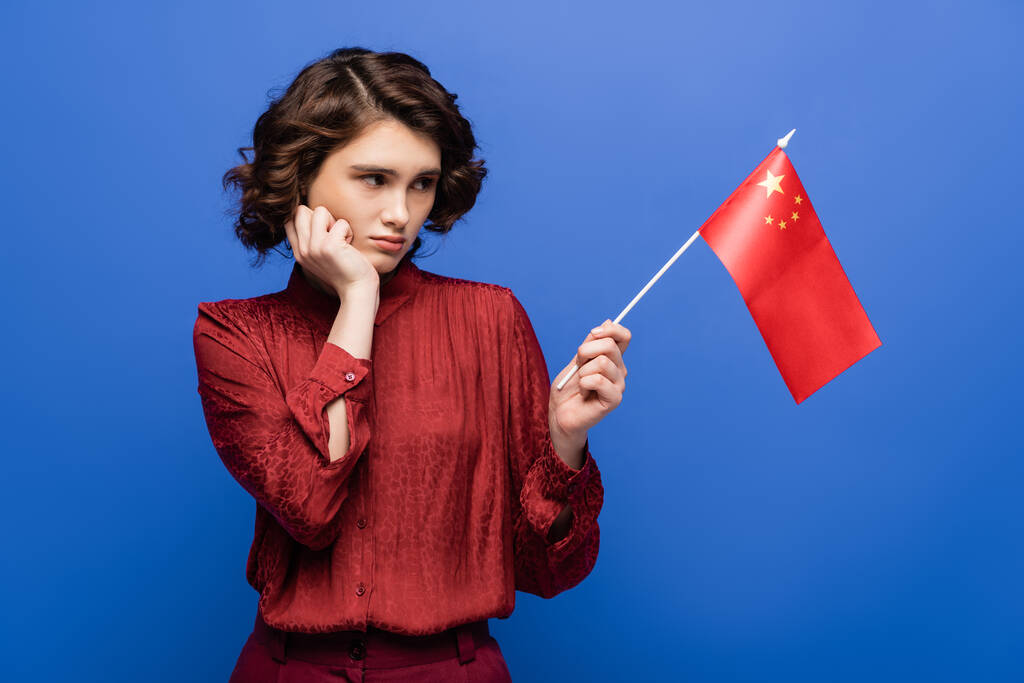 estudiante disgustado con el pelo rizado mirando la bandera de China aislado en azul  - Foto, imagen
