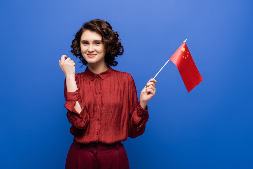feliz joven estudiante con el pelo rizado mirando la bandera de China aislado en azul  - Foto, Imagen