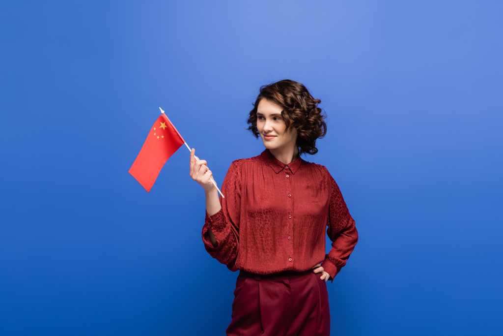 šťastný učitel jazyka drží vlajku Číny, zatímco stojí s rukou na boku izolované na modré  - Fotografie, Obrázek