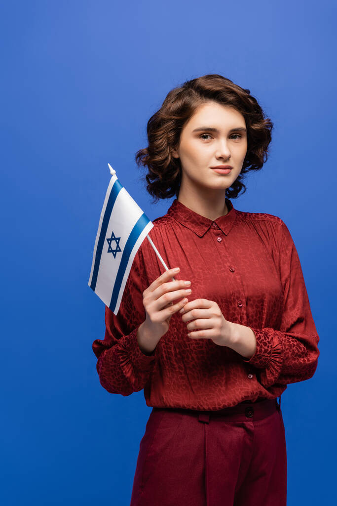 mladý a seriózní učitel jazyků s vlajkou Izraele při pohledu na kameru izolované na modré - Fotografie, Obrázek
