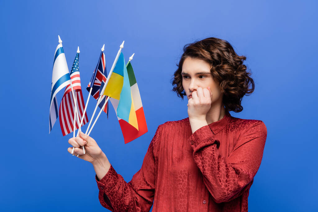 thoughtful woman holding hand near face while looking at flags of different countries isolated on blue - Photo, Image