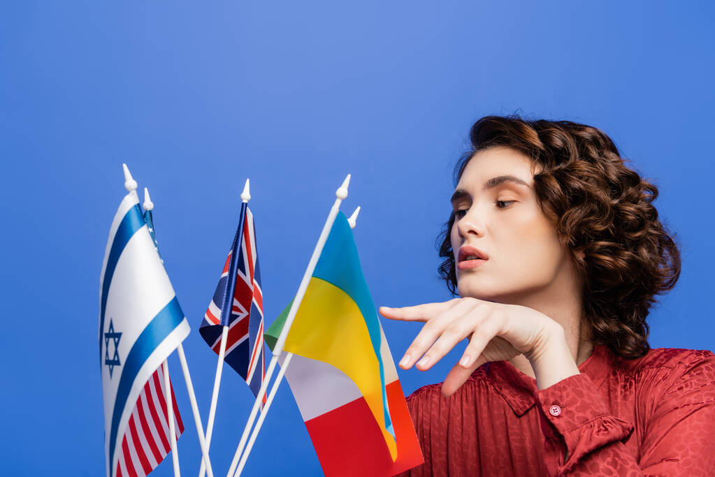 bruneta žena ukazuje na různé vlajky při výběru jazyka ke studiu izolované na modré - Fotografie, Obrázek