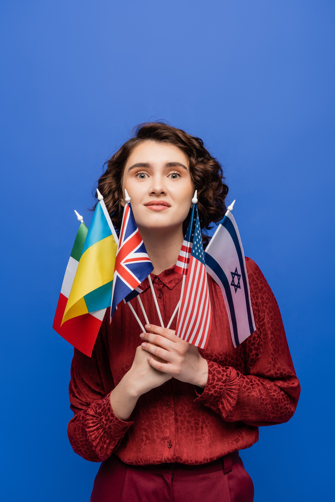 κατάπληκτη γυναίκα κρατώντας σημαίες από διάφορες χώρες και κοιτάζοντας κάμερα απομονωμένη σε μπλε - Φωτογραφία, εικόνα