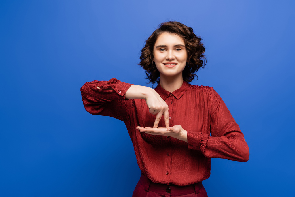 brunette vrouw glimlachen en tonen gebaar betekenis staan op gebarentaal geïsoleerd op blauw - Foto, afbeelding